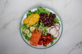 bowl of salad with vegetables ,fresh green oak salad, tomatoes, corn, Red beans, Carrot , red radish, olive oil and cucumber on white marble table. Top view. Food Concept for healthy diet