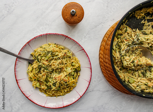 Orzo primavera with green veggies in vintage place over white marble table. One pot pasta with serving spoon, pepper grinder and napkin on set table. 