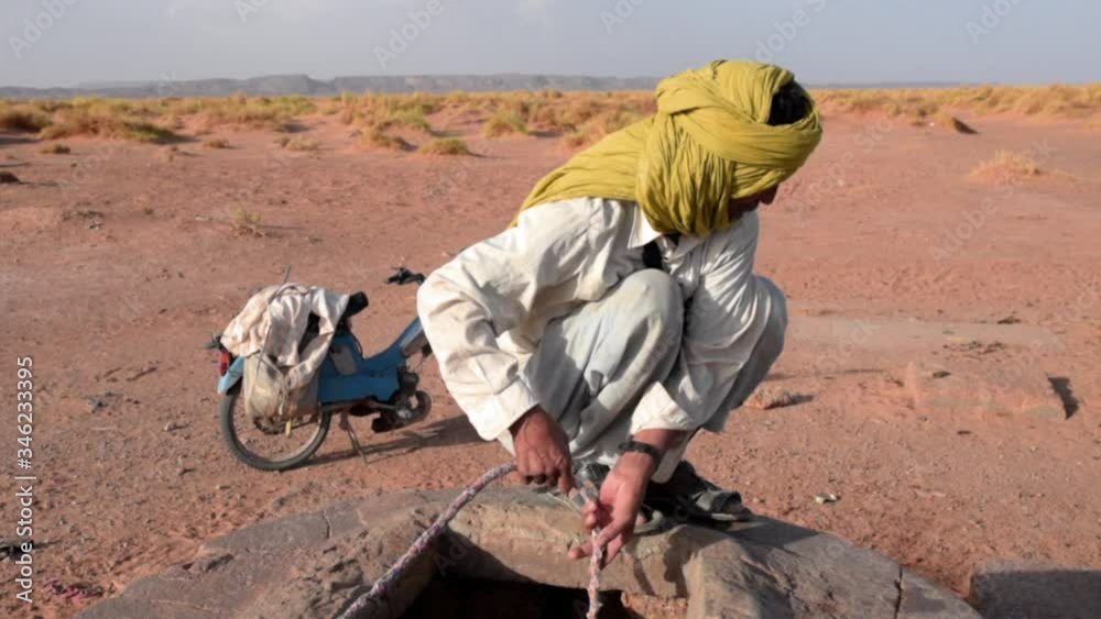 Old man with white clothing and yellow head scarf trying to fill water into the well in the middle of desert in Morocco Old man with white clothing and yellow head scarf trying to fill water into the well in the middle of desert in Morocco