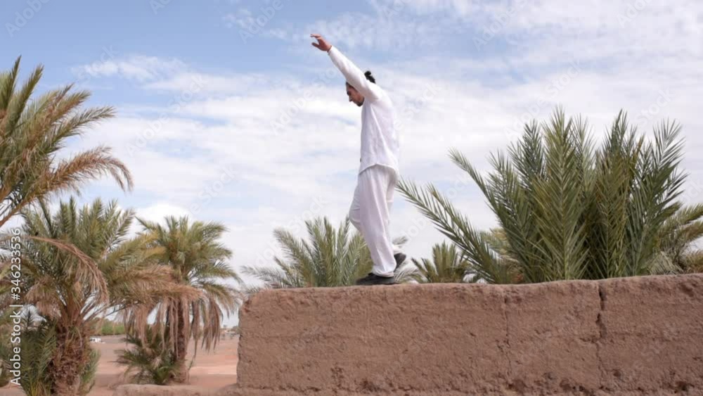 Agile guy exercises balance walking with open arms on a wall of ancient buildings surrounded by desert and palm trees in Morocco Agile guy exercises balance walking with open arms on a wall of ancient buildings surrounded by desert and palm trees in Morocco