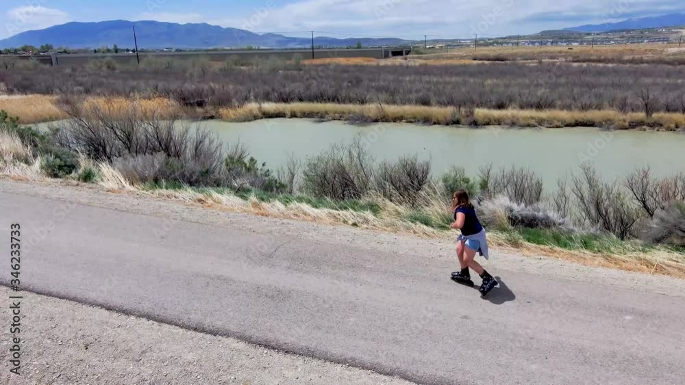 Orbiting a teen girl while she skates along a nature trail beside a river with mountains in the distance Orbiting a teen girl while she skates along a nature trail beside a river with mountains in the distance