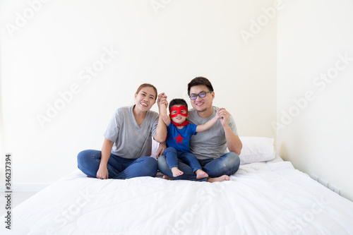 Asian family and son wearing superhero suit hand up and sitting on white bed in the bedroom