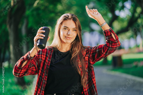 Modern trendy girl listening to music by wireless portable speaker.Young beautiful american woman enjoying,dancing in park. photo