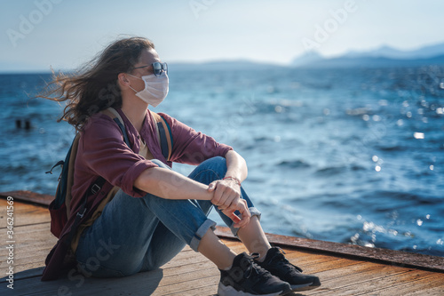 Young girl woman in a mask and sunglasses sits on a wooden pier near the sea. Coronavirus pandemic, weakening of protective measures