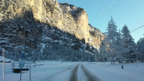 The narrow snow-covered road stretching through the winter forest to the Pederu mountain refuge. Pine trees stand tall and straight. Sun shining on the mountain tops. Forest is already in shadow. photo