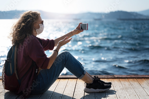 Fotografie One young girl a woman in a mask and sunglasses sits on a wooden pier on the seashore with a smartphone in her hands