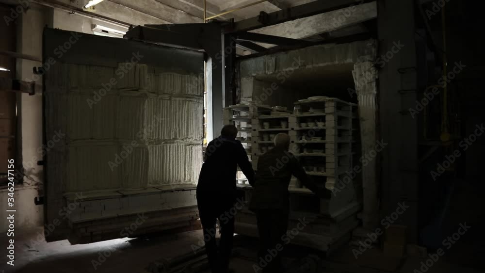 Cookware firing shop. Two men push a mobile rack of earthenware into the oven from left to right. General plan. Grayscale. Cookware firing shop. Two men push a mobile rack of earthenware into the oven from left to right. General plan. Grayscale.