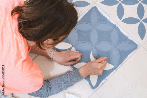 Stay at home and home improvement concept: Close up and top view of a woman holding a brush to paint a decorative template on the floor tiles into gray by using a vintage pattern stencil
