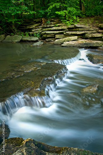 A stream spills over a ledge riffle as it flows through a summer landscape.