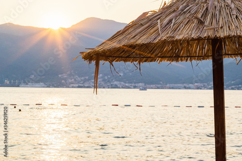 Straw beach umbrella on beach in Montenegro and beautiful sunset behind mountains