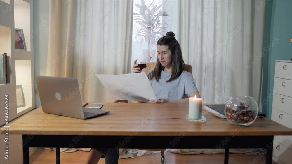 A woman works sitting at a table at home The girl is holding piece of paper and drawing. Woman drinks wine and relax. Remote work. Home office. Self-isolation. Quarantine. Epidemic. Silent evening. A woman works sitting at a table at home The girl is holding piece of paper and drawing. Woman drinks wine and relax. Remote work. Home office. Self-isolation. Quarantine. Epidemic. Silent evening.