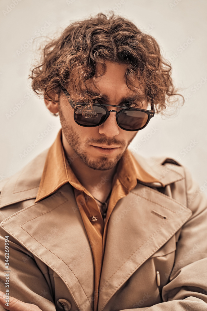 Curly and serious caucasian man standing in a bright studio on a white background, wearing casual and stylish trench coat and sunglasses Curly and serious caucasian man standing in a bright studio on a white background, wearing casual and stylish trench coat and sunglasses