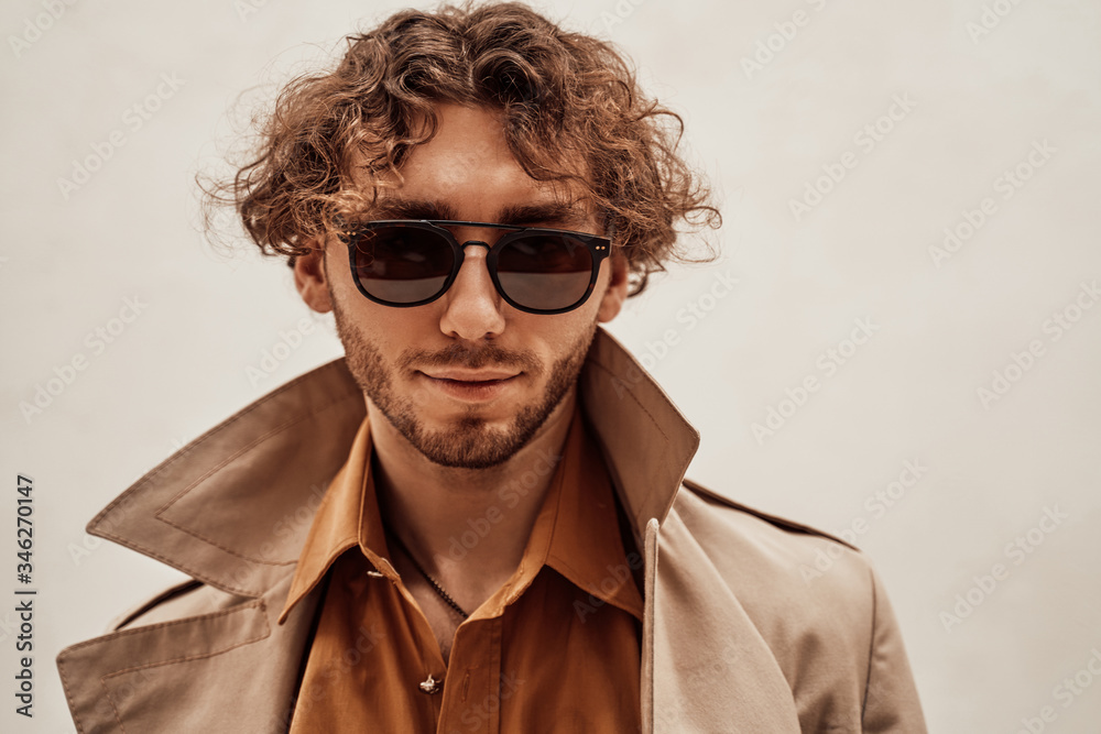 Thoughtful and handsome young man posing in a bright studio on a white background, looking relaxed while wearing trench coat and glasses Thoughtful and handsome young man posing in a bright studio on a white background, looking relaxed while wearing trench coat and glasses