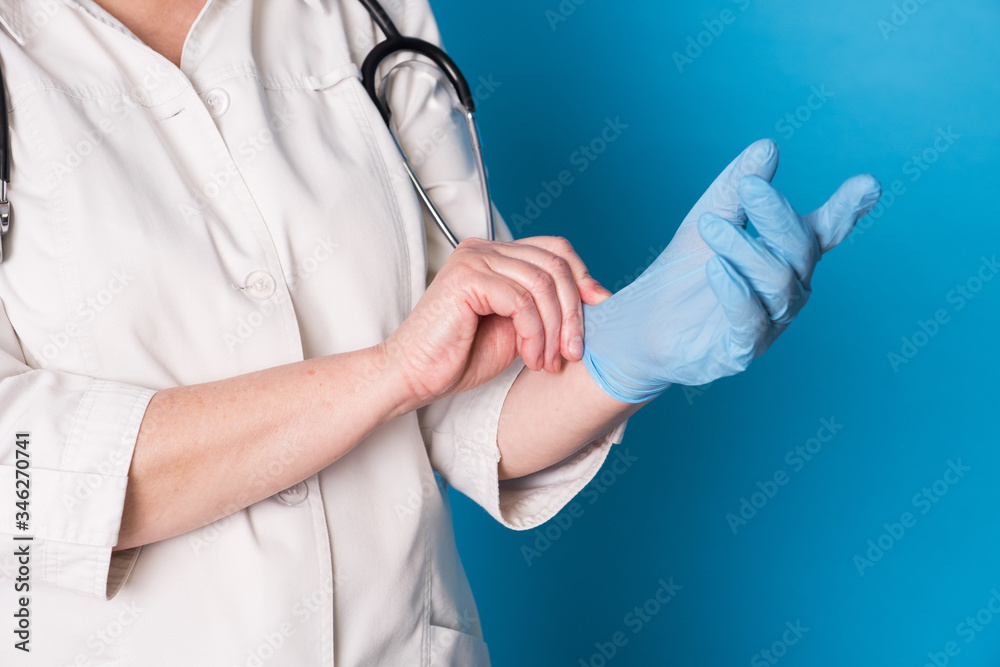 Eldery caucasian doctor lady in white coat and with stethoscope on blue background. Close up of putting on sterille gloves