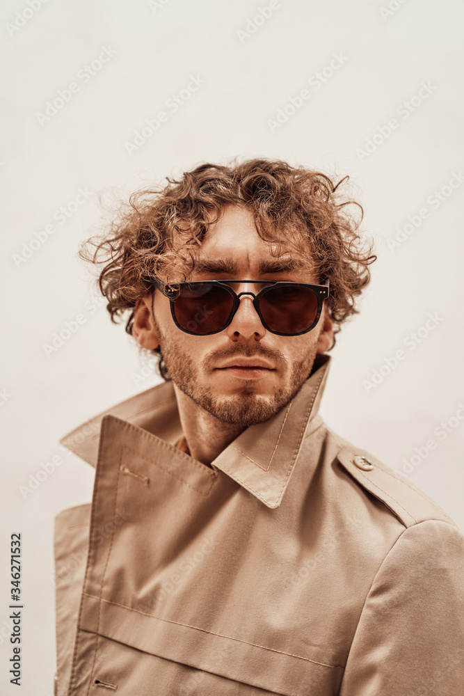 Obraz premium Portrait of a serious young adult guy with curly hair, wearing luxury trench coat, orange shirt and sunglasses, looking authoritative