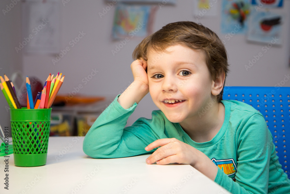 A blond preschooler sits at a white table with colored pencils in a green glass in his room and smiles into the frame. Draw houses during the period of isolation. A blond preschooler sits at a white table with colored pencils in a green glass in his room and smiles into the frame. Draw houses during the period of isolation.