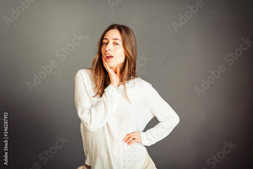 Young smiling girl in a white shirt, head tilted to the side, hand near the cheek, the second in the pocket, insulator on a gray background. No retouching. Without makeup.
