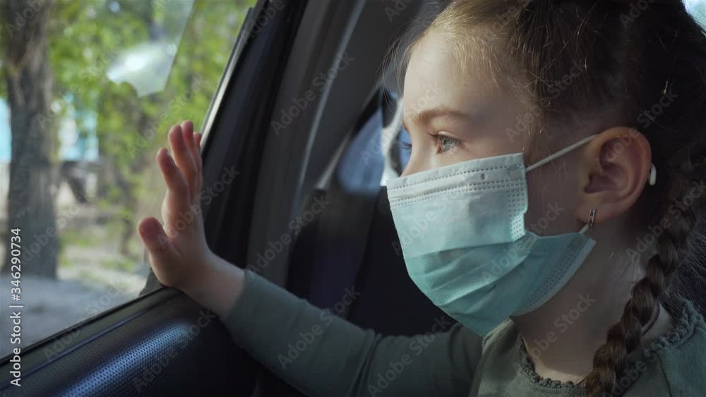Portrait young kid girl in medical mask looks out moving car window on street. Self-isolation in quarantine. Stop COVID-19 infection and pandemic Portrait young kid girl in medical mask looks out moving car window on street. Self-isolation in quarantine. Stop COVID-19 infection and pandemic