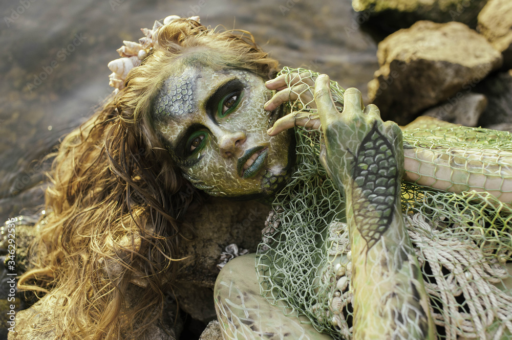 Obraz premium girl mermaid amphibian with green skin with body art with a necklace and a seashell swimsuit around her neck holding her hand to her serious face in the river summer in the water in the lake