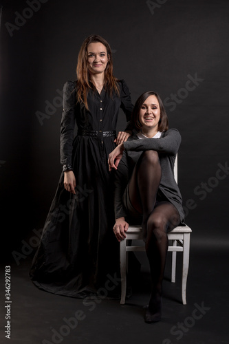 Two makeup girls in dark clothes nearby. One is sitting on a chair, the other is standing behind. Smiling at the viewer. Studio portrait on a black background. Vertical orientation.