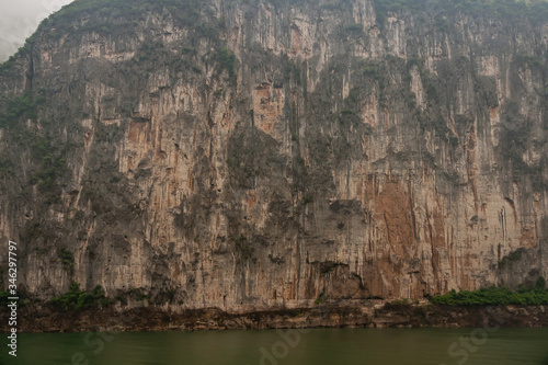 Baidicheng, China - May 7, 2010: Qutang Gorge on Yangtze River. Straigth down brown cliff with some green foliage and darker stalactites above green water.