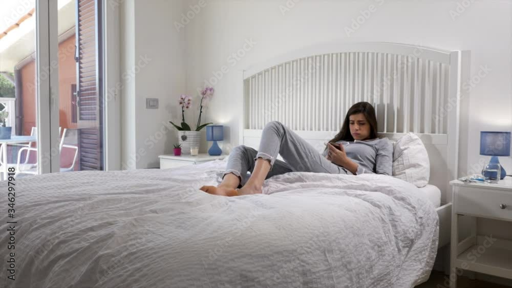 Young woman sitting in bed looking cell phone starting to feel ill Young woman sitting in bed looking cell phone starting to feel ill
