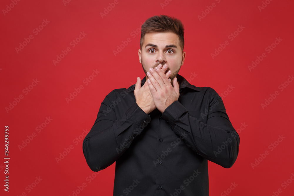 Shocked young bearded guy 20s in classic black shirt posing isolated on red wall background studio portrait. People sincere emotions lifestyle concept. Mock up copy space. Covering mouth with hands. Shocked young bearded guy 20s in classic black shirt posing isolated on red wall background studio portrait. People sincere emotions lifestyle concept. Mock up copy space. Covering mouth with hands.