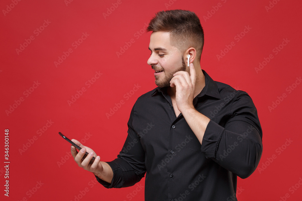 Smiling young bearded guy in classic black shirt posing isolated on bright red wall background studio portrait. People emotions lifestyle concept. Mock up copy space. Using air pods and mobile phone. Smiling young bearded guy in classic black shirt posing isolated on bright red wall background studio portrait. People emotions lifestyle concept. Mock up copy space. Using air pods and mobile phone.
