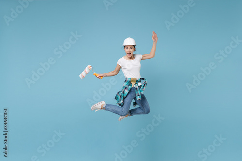 Cheerful young woman in protective helmet hardhat jump with paint roller isolated on blue wall background. Instruments accessories for renovation apartment room. Repair home concept. Spreading hands.