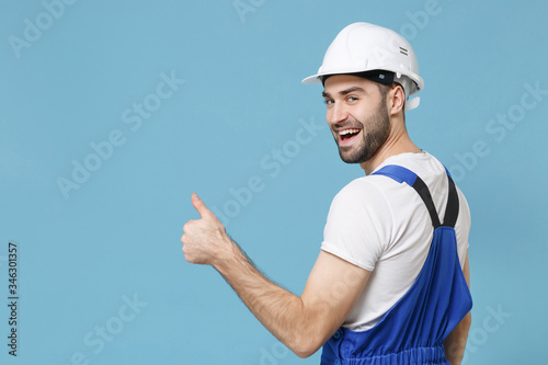 Back rear view of smiling man in coveralls protective hardhat isolated on blue background. Instruments accessories for renovation apartment room. Repair home concept. Looking camera, showing thumb up.