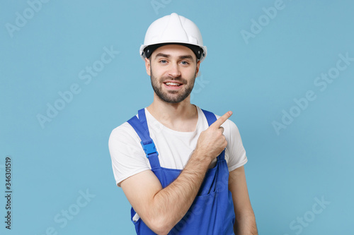 Smiling young man in coveralls protective helmet hardhat isolated on blue wall background. Instruments accessories for renovation apartment room. Repair home concept. Pointing index finger aside up. photo