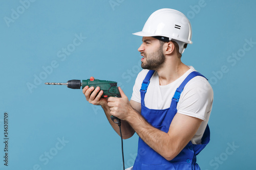 Side view worried young man in coveralls protective helmet hardhat hold electric drill isolated on pastel blue background. Instruments accessories for renovation apartment room. Repair home concept.
