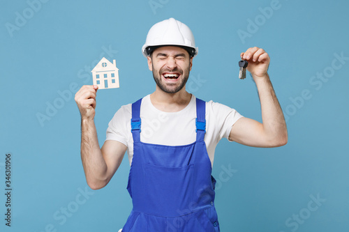 Joyful young man in coveralls protective helmet hardhat hold house bunch of keys isolated on blue wall background in studio. Instruments accessories for renovation apartment room. Repair home concept.