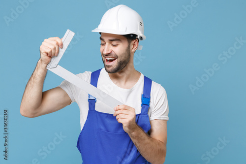 Funny young man in coveralls protective helmet hardhat hold construction building corner isolated on pastel blue background. Instruments accessories for renovation apartment room. Repair home concept.