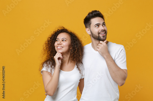 Smiling pensive couple two friends european guy african american girl in white t-shirts posing isolated on yellow background. People lifestyle concept. Put hands prop up on chin, looking aside up.