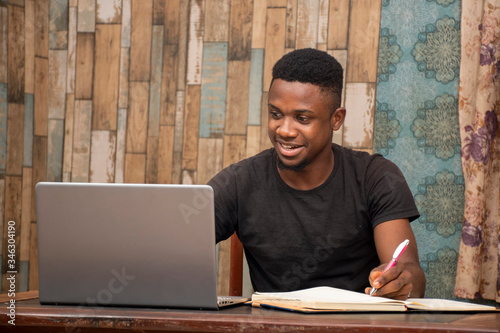 young african man studying at home using his laptop, receiving lectures online and taking notes, smiling