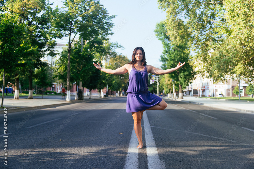 a girl in a lilac dress is doing yoga in the middle of the street