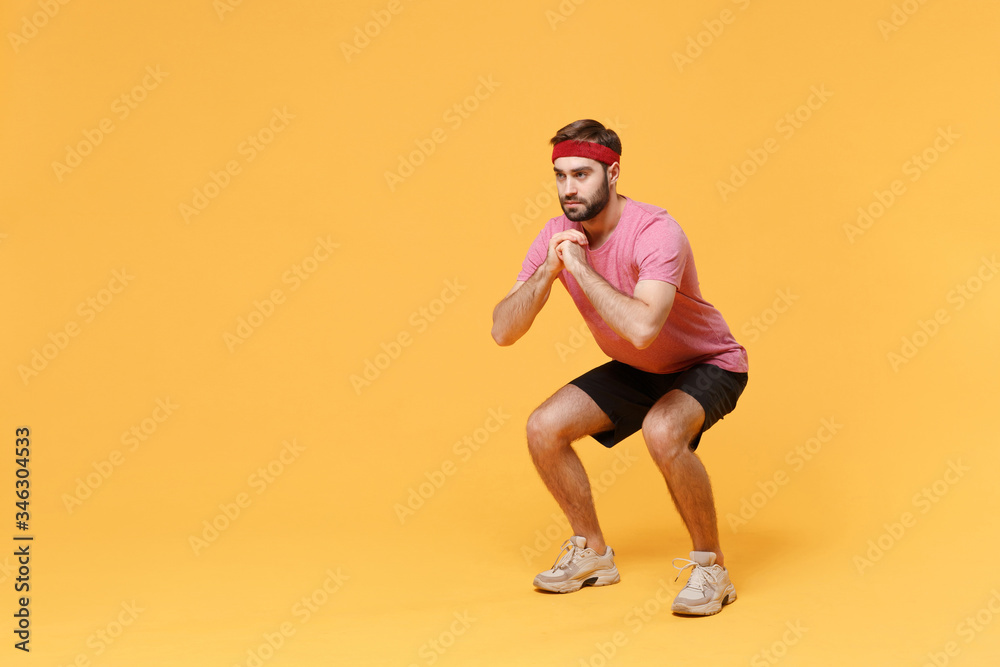 Young bearded fitness sporty guy sportsman in headband t-shirt spend weekend in home gym isolated on yellow background. Workout sport motivation concept. Doing squats exercising, holding hands folded.