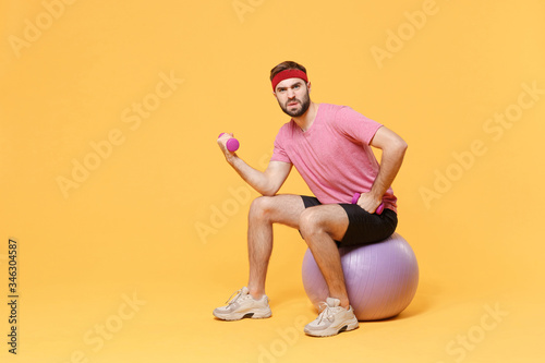 Tensed bearded fitness sporty guy 20s sportsman in headband t-shirt in home gym isolated on yellow background. Workout sport motivation lifestyle concept. Sit on fitball doing exercise with dumbbell.