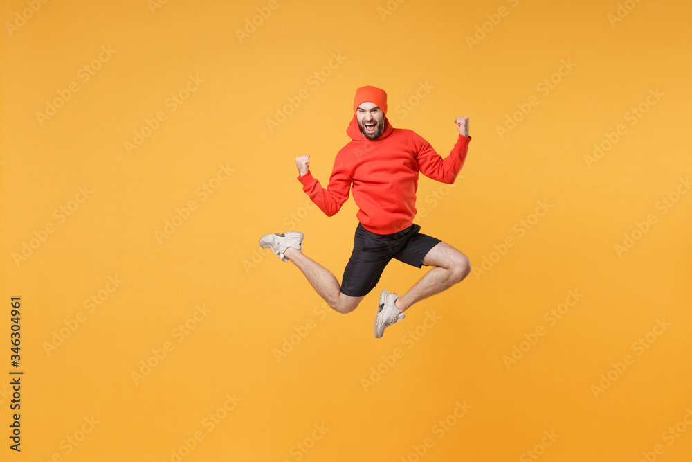 Excited young bearded fitness sporty guy 20s sportsman in hat hoodie shorts in home gym isolated on yellow background. Workout sport motivation lifestyle concept. Jump doing winner gesture screaming.