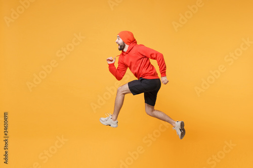 Side view of young bearded fitness sporty guy 20s sportsman in hat hoodie, shorts in home gym isolated on yellow background in studio. Workout sport motivation lifestyle concept. Jumping like running.
