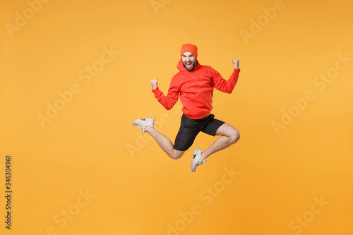 Excited young bearded fitness sporty guy 20s sportsman in hat hoodie shorts in home gym isolated on yellow background. Workout sport motivation lifestyle concept. Jump doing winner gesture screaming. © ViDi Studio