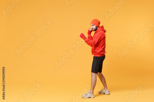 Fototapeta Naklejka Na Ścianę i Meble -  Side view of young bearded fitness sporty guy boxer sportsman in hat, hoodie, shorts in home gym isolated on yellow background. Workout sport motivation concept. Boxing with sports bandages on hands.