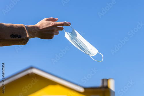 Man takes off medical protective mask and holds it on her finger on blue sky and yellow house on background, enjoys life, clean fresh air after Covid-19 pandemic, self-isolation. Quarantine is over