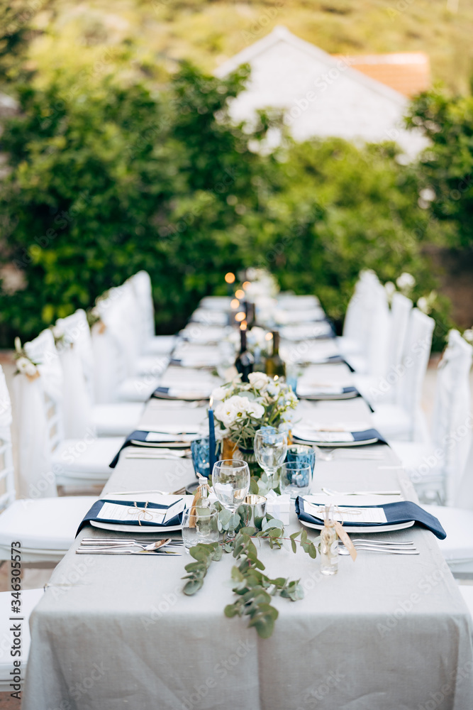 Wedding dinner table reception. Table serving the table with grey tablecloth with white plates, with blue napkins. Flower composition with eucalyptus in the center of the table. White Chiavari chairs