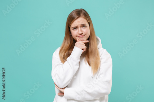 Confused young woman girl in casual white hoodie posing isolated on blue turquoise background studio portrait. People sincere emotions lifestyle concept. Mock up copy space. Put hand prop up on chin.