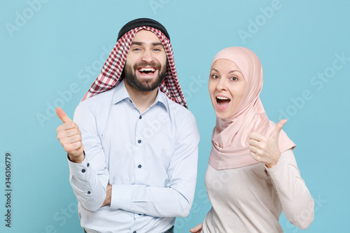 Cheerful young couple friends arabian muslim man wonam in keffiyeh kafiya ring igal agal hijab clothes isolated on blue background in studio. People religious lifestyle concept. Showing thumbs up. photo
