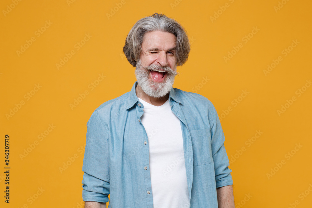 Cheerful elderly gray-haired mustache bearded man in casual blue shirt posing isolated on yellow background studio portrait. People sincere emotions lifestyle concept. Mock up copy space. Blinking. Cheerful elderly gray-haired mustache bearded man in casual blue shirt posing isolated on yellow background studio portrait. People sincere emotions lifestyle concept. Mock up copy space. Blinking.
