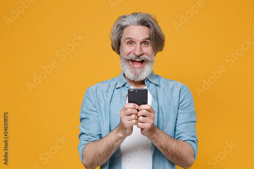 Excited elderly gray-haired mustache bearded man in casual blue shirt posing isolated on yellow background studio. People lifestyle concept. Mock up copy space. Using mobile phone, typing sms message.