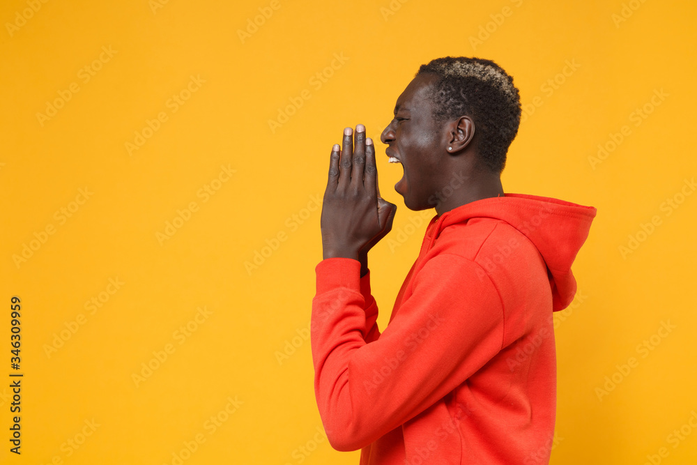 Side view of young african american man guy in red streetwear hoodie posing isolated on yellow wall background. People lifestyle concept. Mock up copy space. Screaming with hand gesture near mouth. Side view of young african american man guy in red streetwear hoodie posing isolated on yellow wall background. People lifestyle concept. Mock up copy space. Screaming with hand gesture near mouth.